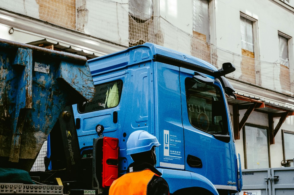 Blue commercial truck at a construction site with a worker in high-visibility clothing and industrial skip nearby